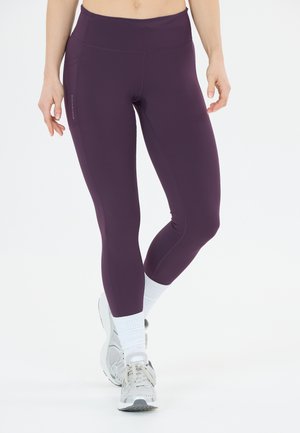 Person iført lilla højtaljede leggings, hvide sokker og hvide sneakers, stående mod en helt hvid baggrund. Overkroppen er ikke fuldt synlig.