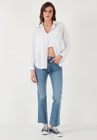 White, long-sleeve button-up shirt over a cropped top, paired with light blue flared jeans and navy sneakers. Subtle pocket detail on shirt.