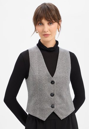 Frau mit braunen Haaren, die goldene Creolen, einen schwarzen Rollkragenpullover und eine graue Fischgrätenweste mit Knöpfen trägt, steht vor weißem Hintergrund.