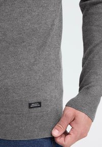 Grå strikket sweater med ribbede manchetter og kant. Har et logo-patch på den nederste side. Stoffet virker blødt og tekstureret.
