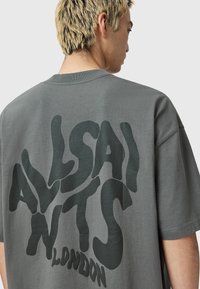 Mężczyzna z krótkimi blond włosami, noszący oversize'owy szary t-shirt z dużym ciemnym napisem "ALLSAINTS LONDON" na plecach.