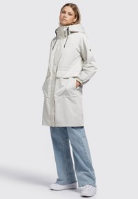 Parka leggera di colore crema con cappuccio, due tasche anteriori, polsini elastici e chiusura con zip; tessuto liscio e traspirante.