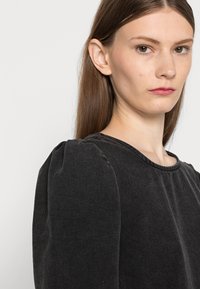 Top preto texturizado de mangas compridas com decote redondo e mangas bufantes, apresentando um discreto detalhamento em costura ao longo do decote.