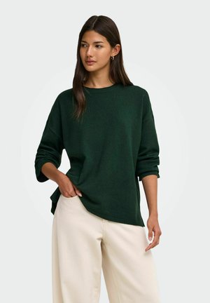 Jeune femme aux longs cheveux foncés portant un pull vert foncé à manches longues et un pantalon ample beige clair, debout avec une main dans la poche.