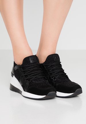 Baskets basses noires avec des accents texturés et pailletés, lacets noirs, semelles intermédiaires blanches, portées pieds nus sur un fond gris uni.