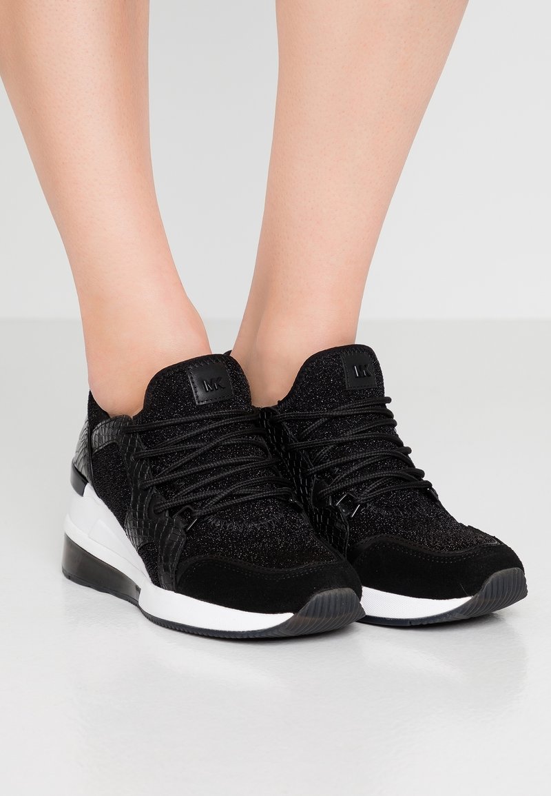 Baskets basses noires avec des accents texturés et pailletés, lacets noirs, semelles intermédiaires blanches, portées pieds nus sur un fond gris uni.