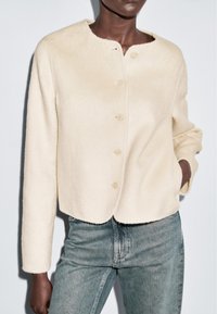 Donkerhuidig persoon in een crèmekleurige, knoopsluiting wollen jas met lange mouwen en lichtblauwe spijkerbroek, met één hand in de zak.