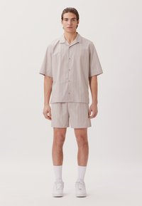 Chemise à manches courtes, boutonnée, et short assorti en tissu beige clair avec des rayures bleues subtiles. Des baskets blanches et des chaussettes de cheville complètent le look.