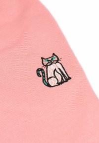 Roze stof met een geborduurd kattendesign in wit en groen. De kat heeft een zittende houding, met puntige oren en een opgerolde staart.