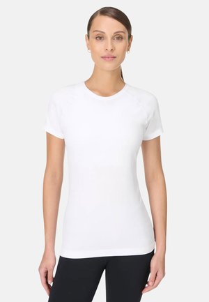 Vrouw met bruin haar in een witte, aansluitende t-shirt met korte mouwen en ronde hals en zwarte broek, staand tegen een effen lichte achtergrond.