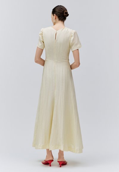 Frau mit zu einem Dutt gebundenem Haar, die ein cremefarbenes Midikleid mit kurzen Puffärmeln und roten Absatzsandalen trägt, steht mit verschränkten Händen hinter dem Rücken.