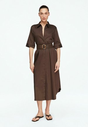 Donna in piedi che indossa un abito midi marrone con bottoni, maniche a tre quarti, una cintura larga intrecciata e sandali infradito neri.