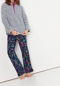 Kobieta w pasiaku polarowym i granatowych dżinsowych spodniach w kwiaty, stoi boso przy białej ścianie.