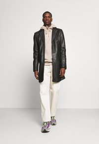 Melna ādas jaka virs bēšas kapuci, baltas bikses un daudzkrāsaini sporta apavi. Gluda tekstūra, pieguļošs dizains un redzami šuvumu elementi.