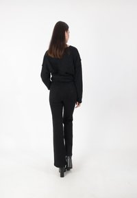 Donna con lunghi capelli castani, che indossa un maglione nero, pantaloni neri e stivali con tacco neri, in piedi di fronte a uno sfondo bianco.