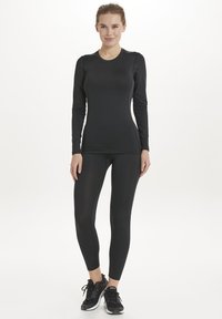 Schwarzes, langärmliges Sportoberteil und enganliegende Leggings, aus dehnbarem Material. Glatte Textur mit taillierter Passform, kombiniert mit schwarzen Sportschuhen.