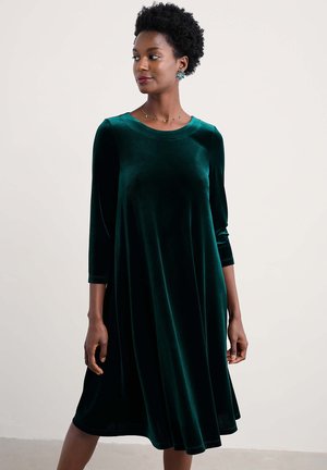 Ein tiefgrünes, knielanges Samtkleid mit drei Viertel-Ärmeln, einem runden Ausschnitt und einer fließenden A-Linie.