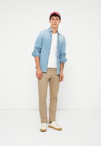 Lys denimskjorte, hvit t-skjorte, khaki-bukser og hvite sneakers med beige såler. Modellen poserer mot en enkel hvit bakgrunn.