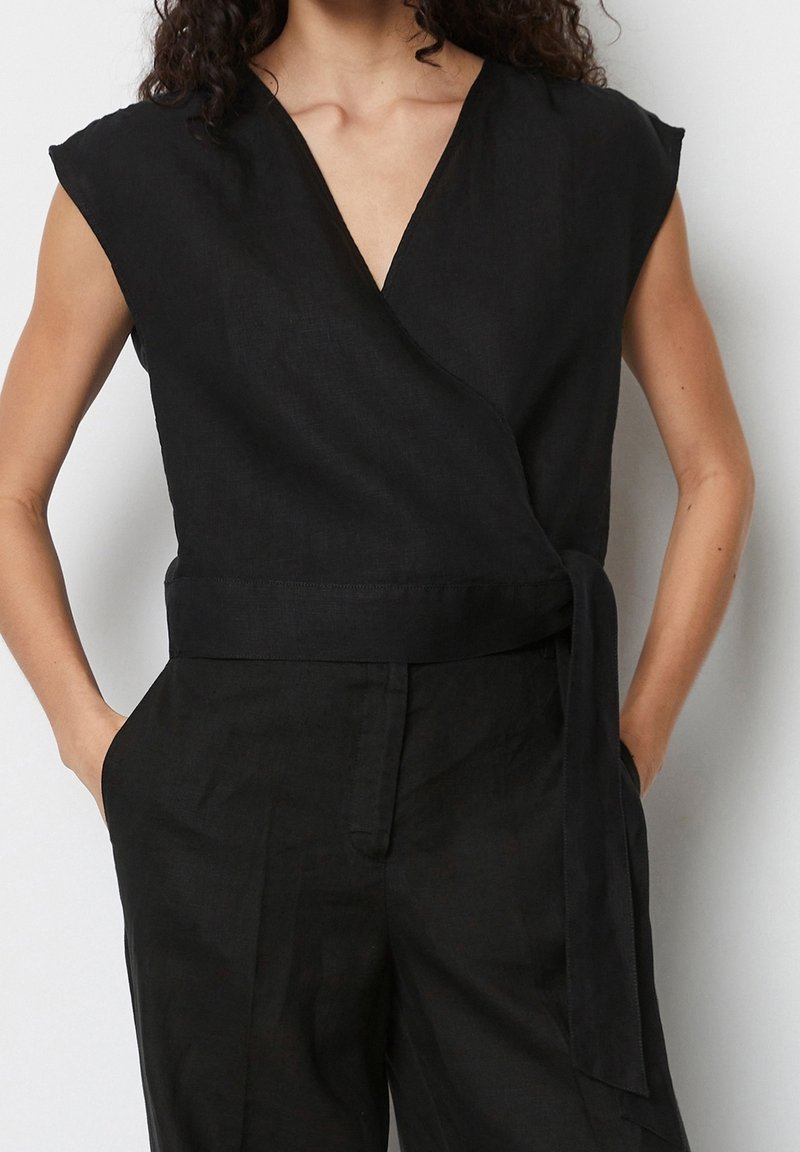 Femme portant un haut cache-cœur noir sans manches et un pantalon noir assorti, mains dans les poches, debout devant un fond blanc.