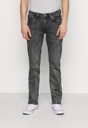 Person wearing faded black slim-fit jeans with a white t-shirt and white sneakers, standing against a plain light background.