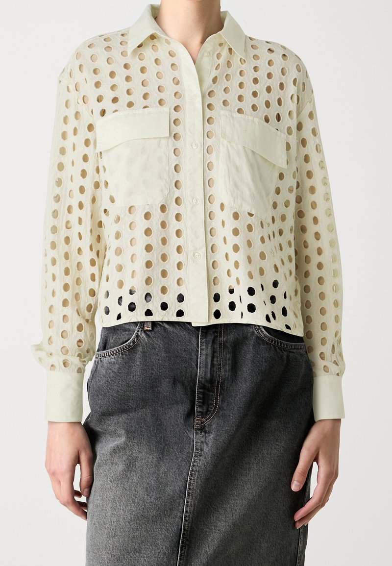 Light beige cropped shirt with large cut-out circles, two chest pockets, and a button-up front. Paired with a dark denim skirt.