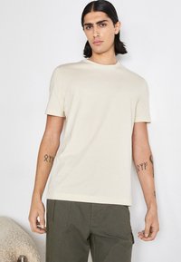 T-shirt beige à manches courtes avec un col rond, fabriqué dans un tissu doux. Présente un branding discret sur la poitrine et une coupe décontractée.