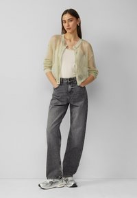 Ein hellgrüner Netz-Cardigan über einem weißen Oberteil, kombiniert mit hochgeschnittenen grauen Jeans und silbernen Sneakers, was einen lässigen Look kreiert.