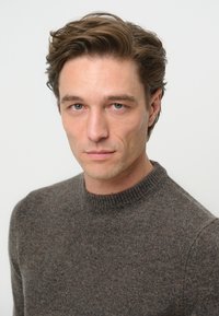 Brun ulltröja med rund hals, med ett strukturerat stickat mönster och en avslappnad passform. Modellen har kort, rufsig hår.