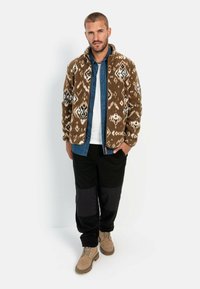 Braune, gemusterte Fleecejacke mit weißen und schwarzen geometrischen Designs, über einem Denimhemd getragen, kombiniert mit schwarzen Hosen und beigen Stiefeln.