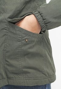 Oliwkowa kurtka z miękkim, teksturowanym wykończeniem, z przednią kieszenią zapinaną na mosiężny zatrzask i elastycznymi mankietami.