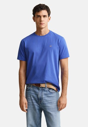 Joven con una camiseta azul lisa de manga corta y vaqueros azul claro con un cinturón beige, de pie frente a un fondo blanco.