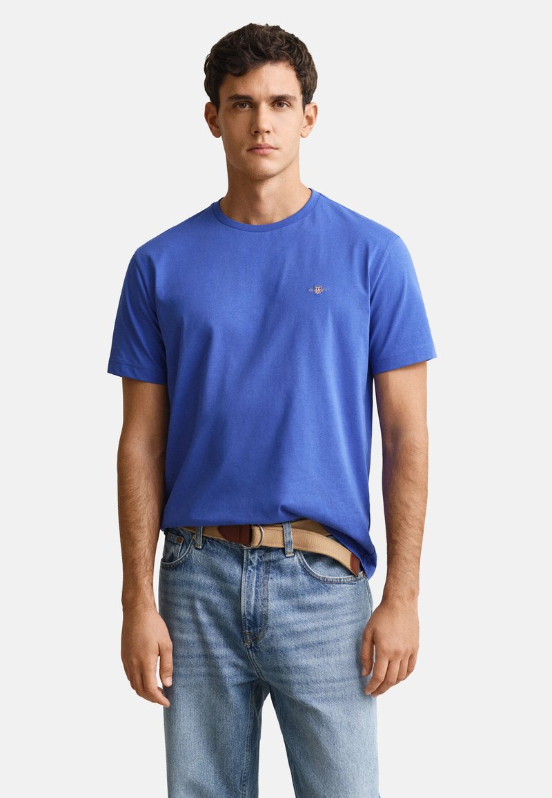 Jeune homme portant un t-shirt bleu uni à manches courtes et un jean bleu clair avec une ceinture beige, debout devant un fond blanc.
