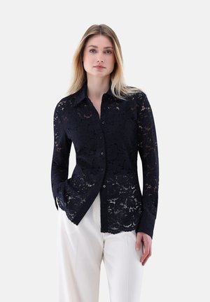 Navy lace shirt with a button-down design, featuring a floral pattern, long sleeves, and a slightly sheer texture. Paired with white pants.