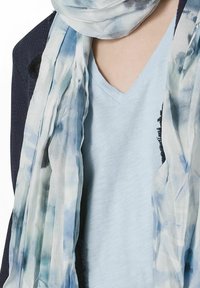 Let vaskelig tørklæde i akvarel blå og hvid, med et blødt materiale, flydende drapering og subtilt abstrakt mønster, lagd over en lys top.