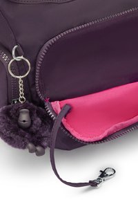 Lilla stofpose med en pink inderside, der har en lynlås, kæde og en fluffy lilla pom-pom nøglering. Sølv hardwaredetaljer inkluderet.