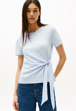 Jasnoniebieski t-shirt z krótkim rękawem z związanym węzłem w okolicy talii. Wykonany z miękkiego materiału, z okrągłym dekoltem i luźnym krojem. W połączeniu z ciemnymi dżinsami.