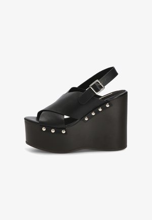 Sandalia de cuña con plataforma negra, tiras cruzadas, hebilla en el tobillo y tachuelas plateadas a lo largo del borde de la suela en un tacón de cuña sólido.