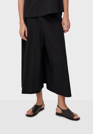 Osoba ubrana w czarne, szerokie, skrócone spodnie, czarny top bez rękawów, czarne sandały na obcasie z odkrytymi palcami oraz srebrny pierścionek na lewej ręce.