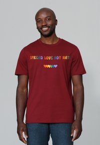 Bordeauxfarbene Baumwoll-T-Shirt mit dem Aufdruck "VERBREITE LIEBE, NICHT HASS" in bunten Buchstaben und einer Reihe bunter Herzformen unter dem Text.