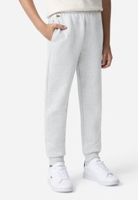 Grå joggebukser med elastisk midjebånd, sidelommer og ribbestrikkede mansjetter. Har en liten grønn Lacoste-logo på venstre hofte.