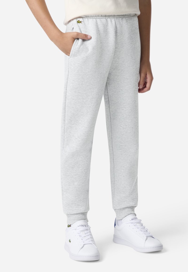 Grå joggebukser med elastisk midjebånd, sidelommer og ribbestrikkede mansjetter. Har en liten grønn Lacoste-logo på venstre hofte.