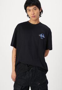 Black cotton t-shirt with short sleeves, featuring a blue and white logo on the left chest. Soft texture with a relaxed fit.