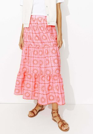 Femme portant une jupe midi rose à volants avec des motifs de soleil orange, un haut blanc et des sandales gladiateur brunes à clous.