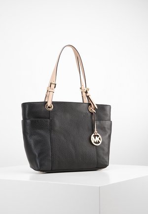 Sac fourre-tout en cuir noir texturé avec doubles anses beige et charm logo MK doré sur le côté, posé sur un socle blanc.