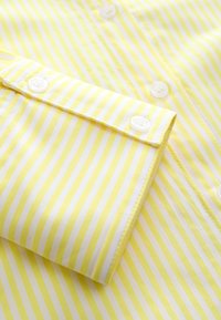 Geltonos ir baltos vertikalios juostelės marškinių rankogalis, užsegamas baltais mygtukais su matomomis siūlėmis.