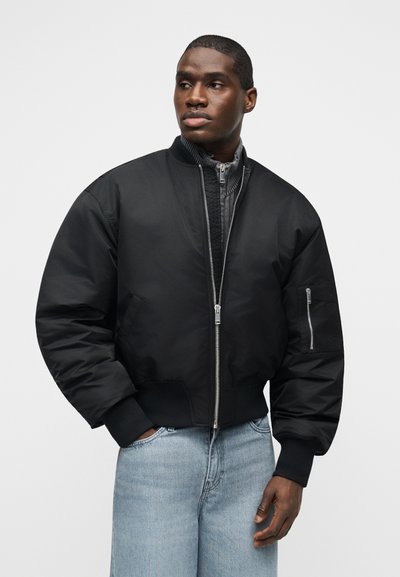 Μαύρο μπουφάν bomber με φερμουάρ μπροστά και φερμουάρ σε τσέπη του μανικιού, φορεμένο πάνω από γκρι τοπ με φερμουάρ και ανοιχτό μπλε τζιν από έναν άνδρα που στέκεται.