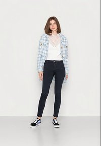Casaco azul xadrez abotoado sobre um top de renda branca, jeans skinny pretos e sapatilhas pretas com detalhes azuis. Tecido suave, design ajustado.
