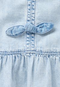 Lys blå denimkjole med detalj av en knyttet sløyfe i midten, rynket falmehank og synlige sømmer.