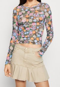 Långärmad, figurnära blus med blommönster i flera färger (rosa, blå, orange) på en mörk bakgrund. Materialet är genomskinligt med scallopade kanter. Matchas med en beige utsvängd kjol.