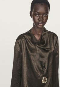 Blusa marrone scuro, texturizzata, con scollo drappeggiato, abbinata a una collana di corda nera con un pendente in argento dalla forma organica.